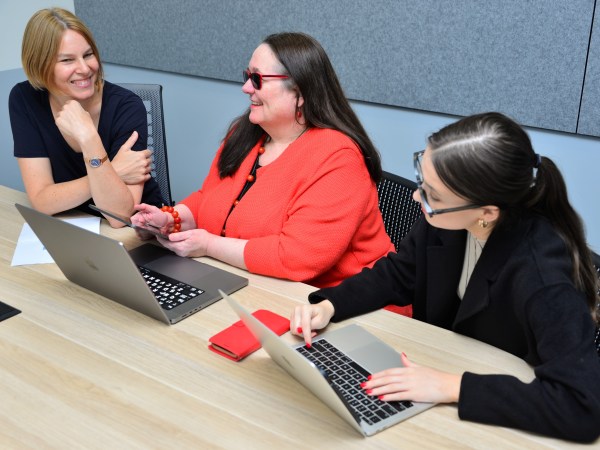 Annmaree Watharow and Monika Bednarek are smiling as they are sitting at a table during a research meeting communicating with the help of a tablet device (for speech-to-text transcription), while accessibility assistant Susannah McNally is using a laptop for additional live transcription.
